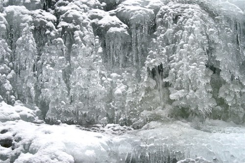 A close-up of a frozen waterfall, with intricate ice formations and glistening icicles against a snowy backdrop.
