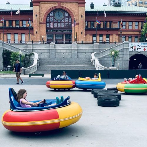 Children enjoy bumper cars in a vibrant outdoor setting near a historic building, with flags and greenery in the background.