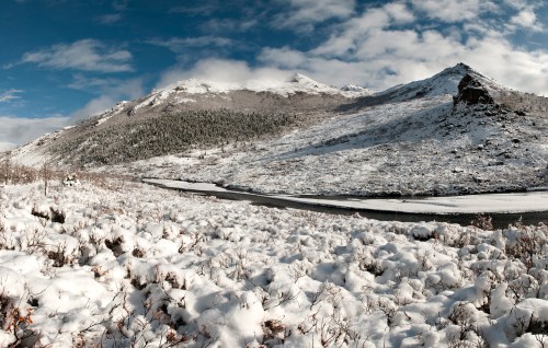 Snow-covered landscape with mountains and a river under a blue sky with scattered clouds.