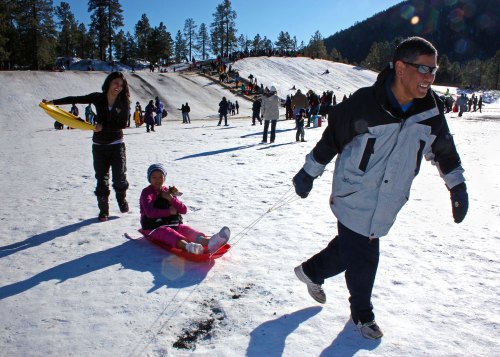 A snowy hill filled with people sledding; a man runs while pulling a girl on a sled, and a woman holds a sled nearby.
