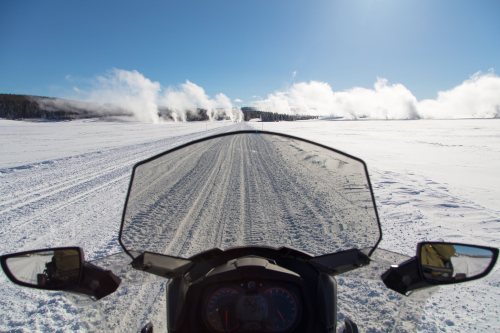 View from a snowmobile on a snowy path, with steam rising from geysers in the distance under a clear blue sky.