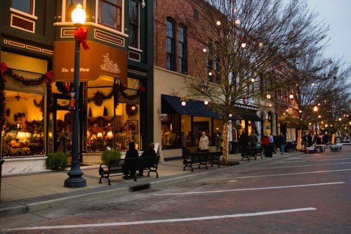 A festive street scene with decorated shops, benches, and people enjoying the holiday atmosphere at dusk.