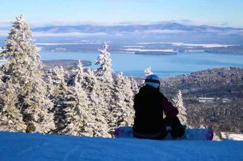 A snowboarder sits on a snowy slope, overlooking a scenic view of a lake and mountains in the distance.