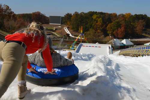 A person pushes a friend on a snow tube down a snowy slope, with amusement park rides in the background.