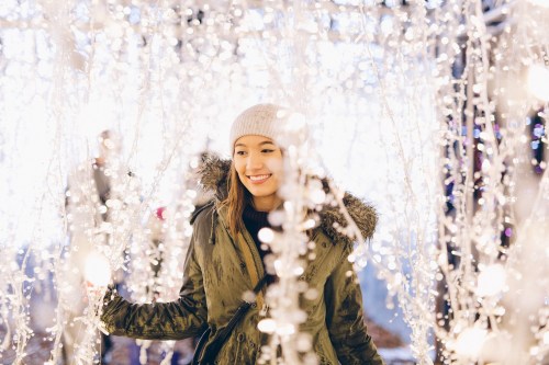 A smiling woman in a cozy coat stands among sparkling lights, creating a festive and joyful atmosphere.