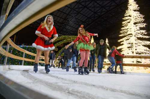 Two women in festive outfits skate on an ice rink, surrounded by holiday lights and a decorated Christmas tree.