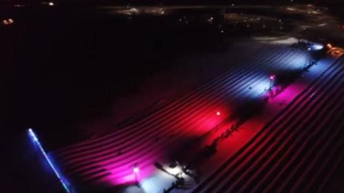 Aerial view of a snow-covered field illuminated by colorful lights in pink and blue, with shadows of people visible.