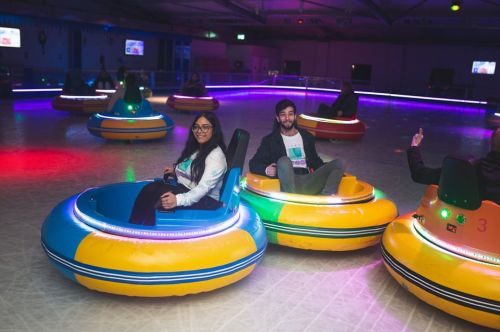 Two people smile while sitting in colorful bumper cars on a rink, surrounded by others and vibrant lights.