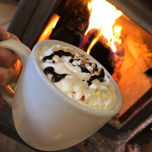 A cozy mug of hot chocolate topped with whipped cream and chocolate drizzle, with a fireplace glowing in the background.