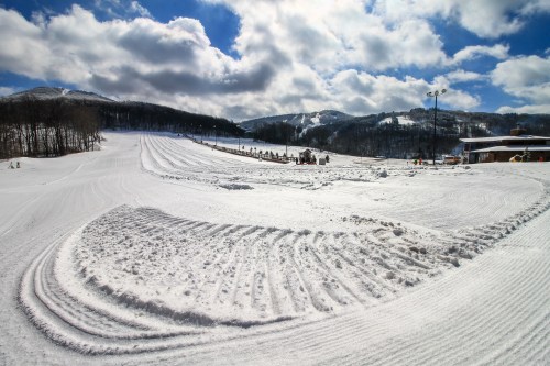 A snowy landscape with groomed ski trails, mountains in the background, and a partly cloudy sky.