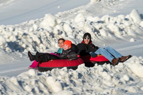 Three people enjoy tubing down a snowy hill, smiling and wearing winter clothing, with snow mounds in the background.