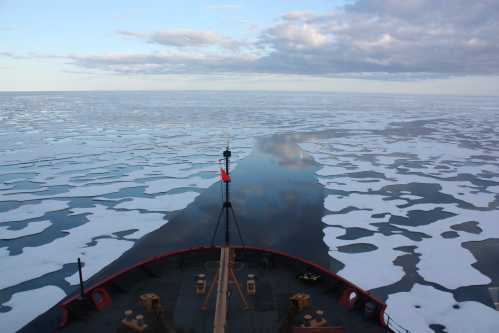 View from the bow of a ship navigating through icy waters, with patches of ice and a cloudy sky above.