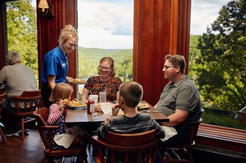 A waitress serves food to a family at a table with a scenic view of nature in a cozy restaurant.