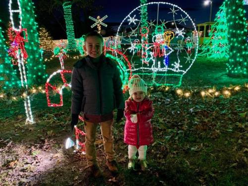 Two children stand in front of colorful holiday lights, smiling in winter clothing amidst festive decorations.