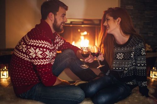 A couple in festive sweaters smiles at each other while holding glasses of wine by a cozy fireplace.