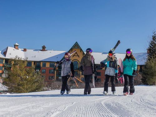 Four skiers walking on a snowy path with a lodge in the background, wearing winter gear and colorful goggles.