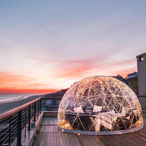 A cozy transparent dome on a deck overlooking a beach at sunset, with vibrant colors in the sky.