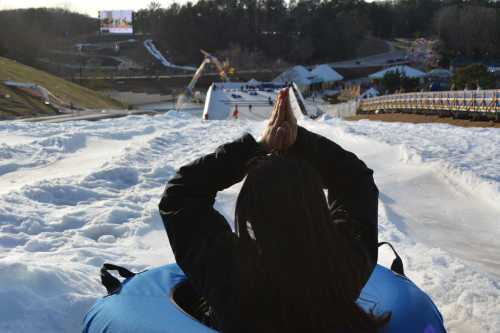 A person sits in a blue snow tube, hands together, looking down a snowy slope with a large screen in the background.