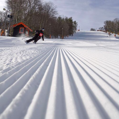 A skier glides down a snowy slope, with freshly groomed tracks and trees in the background.
