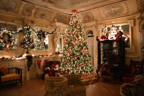 A beautifully decorated room with a large Christmas tree, festive garlands, and cozy seating, all illuminated by warm lights.