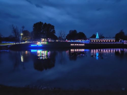 A serene night scene with colorful lights reflecting on a calm lake, surrounded by trees and festive decorations.