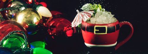 A festive drink in a Santa-themed cup, garnished with mint and an umbrella, surrounded by colorful holiday ornaments.
