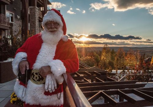 Santa Claus in a red suit stands on a balcony, smiling against a sunset backdrop with colorful autumn trees.