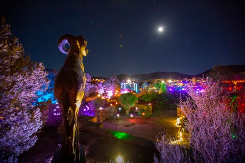 A statue of a ram overlooks a colorful, illuminated landscape under a night sky with a bright moon.