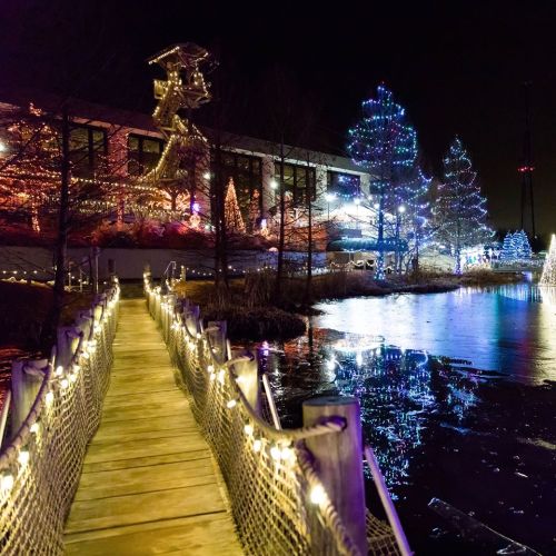 A lit pathway leads to a building adorned with colorful lights, surrounded by trees and a reflective water surface.