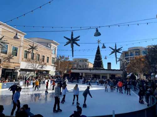 A bustling outdoor ice skating rink surrounded by festive decorations and a large Christmas tree under a clear blue sky.