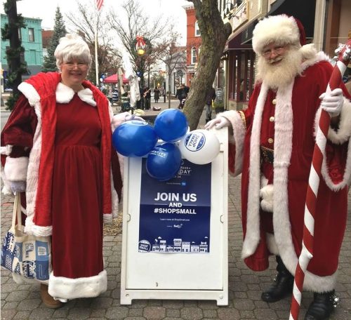A woman in a festive red outfit and Santa Claus pose next to a sign promoting small business shopping.