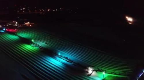 Aerial view of a field at night, illuminated by colorful lights in green, blue, and red, with vehicles visible.
