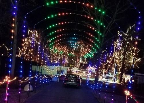 A car drives through a colorful archway of holiday lights in a festive, illuminated park at night.