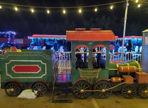 A colorful, decorated train ride with festive lights, set against a night sky. People are enjoying the attraction nearby.