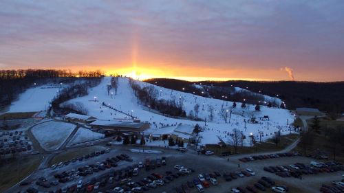 A snowy hill at sunset, with skiers and snowboarders visible, and a parking lot filled with cars in the foreground.