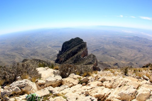 A panoramic view from a mountain peak, showcasing rugged terrain and a distant horizon under a clear blue sky.