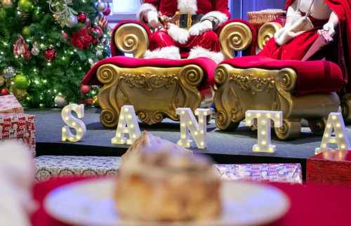 Santa and Mrs. Claus sit on ornate chairs beside a decorated Christmas tree, with "SANTA" in lights and a dessert in the foreground.