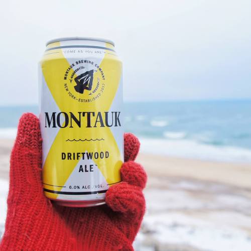 A person in red gloves holds a can of Montauk Driftwood Ale by the beach, with ocean waves in the background.