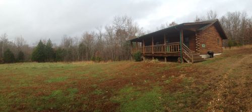 A rustic wooden cabin surrounded by a grassy field and trees under a cloudy sky.