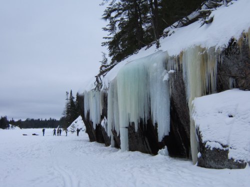 Icicles hang from a rocky cliff in a snowy landscape, with silhouettes of people in the background.