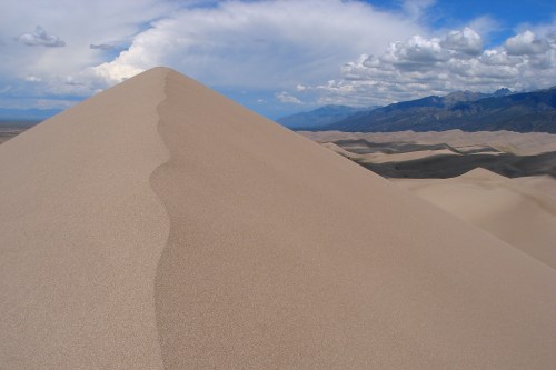 A sandy dune rises sharply against a backdrop of blue sky and distant mountains, with fluffy clouds above.