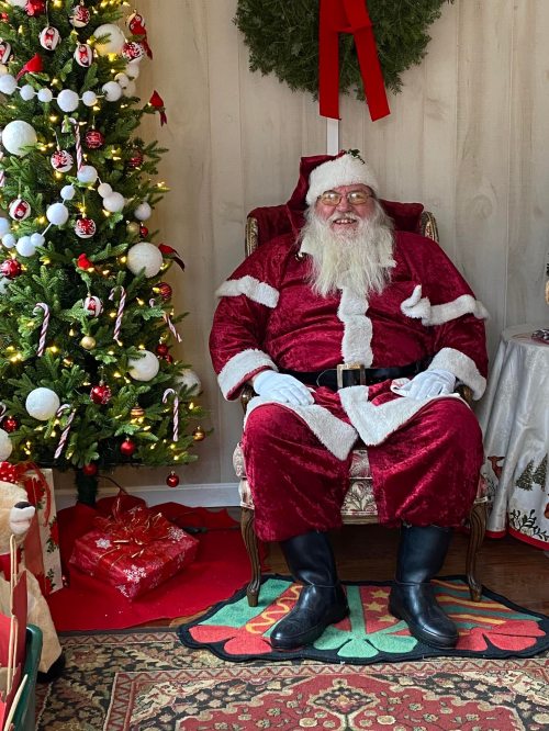Santa Claus in a red suit sits in a cozy room decorated for Christmas, with a tree and gifts nearby.