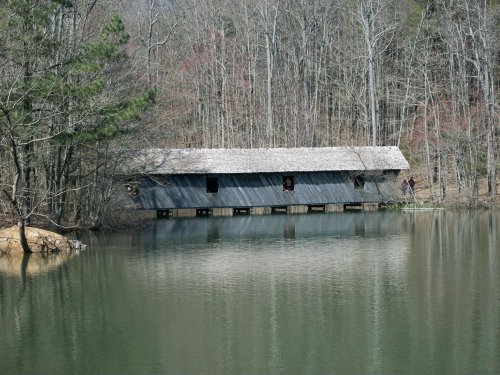 A rustic covered bridge over a calm lake, surrounded by trees in early spring.