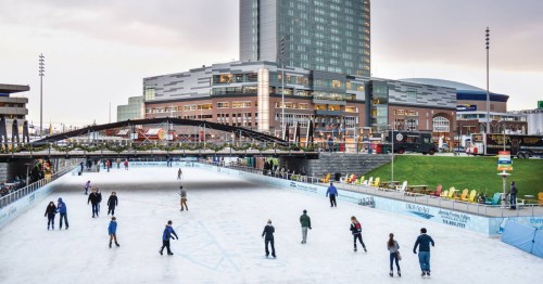 People ice skating on a rink with a city skyline and buildings in the background.