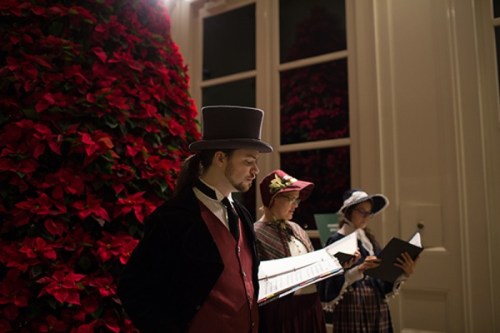 Three carolers in period costumes sing near a large poinsettia display, with one wearing a top hat.