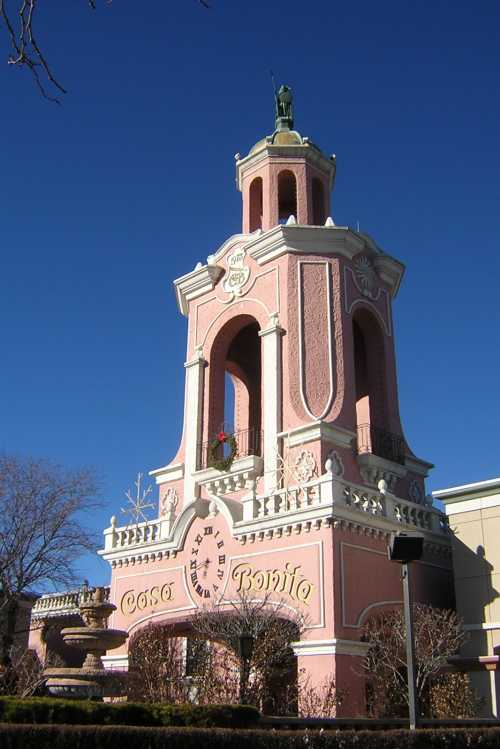 A tall, pink building with a clock and decorative tower, featuring a statue on top against a clear blue sky.