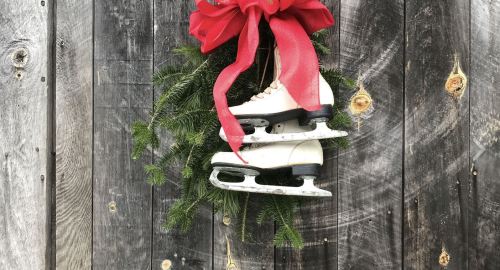 A pair of white ice skates adorned with a red bow, hanging on a wooden wall with evergreen branches.