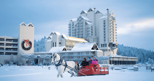 A horse-drawn sleigh in snow, with two people riding, in front of a large hotel decorated for the holidays.