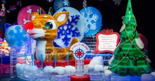 Colorful ice sculpture of a reindeer with a red nose, surrounded by snowflakes and a decorated Christmas tree.