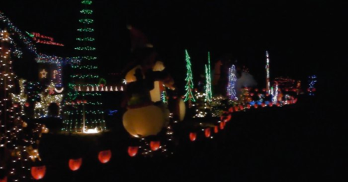 A festive display of colorful Christmas lights and decorations along a dark street, featuring trees and inflatable figures.